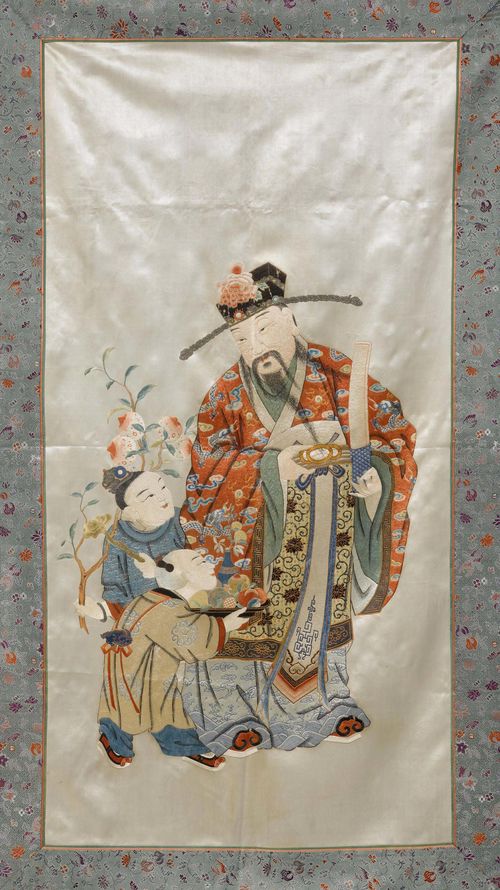 A POLYCHROME SILK EMBROIDERY OF FU, THE GOD OF LUCK, WITH TWO CHILDREN ON A WHITE GROUND WITHIN A BORDER OF FLOWERS AND PEACHES. China, late Qing Dynasty, 130x75 cm.