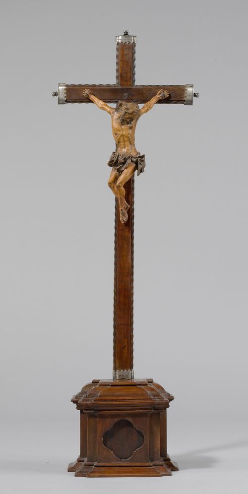 CRUCIFIX,Spain, beginning of the 18th century. Boxwood, carved all-around and patinated. Three nails, Christ with a twisted posture, his legs turned to the side. H 24 cm. The arms of the cross with silver ends. On a later, stepped plinth. H in total 74 cm. Fingers missing.