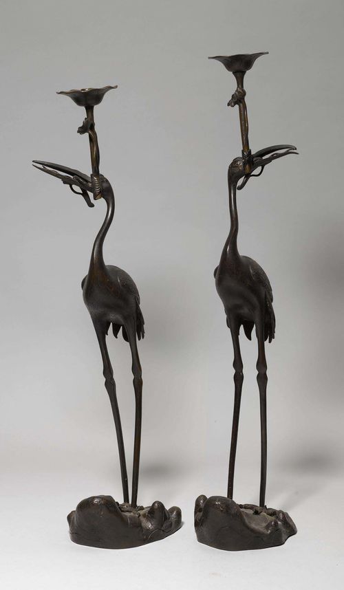 PAIR OF CANDLESTICKS IN THE FORM OF CRANES.Japan, Meiji Period, H 89.5 und 96.5 cm. Bronze. The slender birds stand on rocks, each holding a lotus flower in its beak, which serves as a candle holder. Incised plumage. (2)