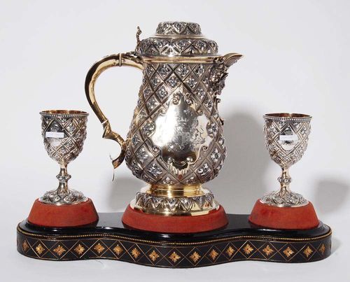 SILVER-GILT SET. London, 1862/63.Maker's mark R. Hennel. Pot and two cups with matching, exquisitely carved wooden tray, velvet lining. Lozenge-shaped decor all around with blossoms. Pot with curved handle, hinged lid, spout mascaron. Over round foot with a zigzag leaf frieze. Cups with a beading finish on the upper rim. In the central cartouches: engraved, crowned coat-of-arms, flanked by a dragon and a lion. H pot 37.5 cm. H cups 18.5 cm. Total weight 3730 g.