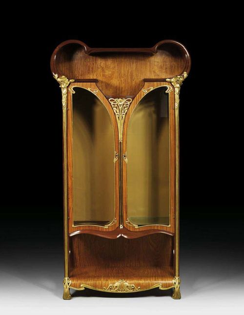 SHAPED VITRINE, probably France, circa 1900. Lemonwood. With rich and finely crafted gilt bronze mounts in the form of poppies. The cornice cracked on both sides. 105x38x200 cm.