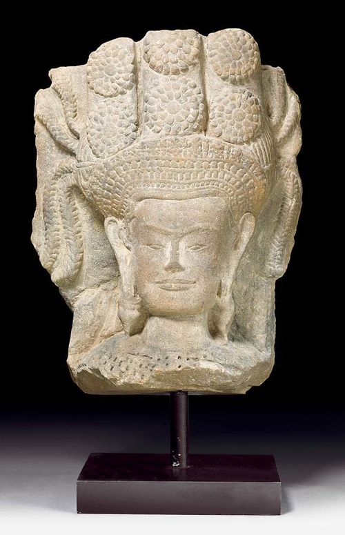 LARGE APSARA HEAD.Khmer, in the style of Angkor Vat, 1st half of the 12th century. H 42 cm. Beige sandstone. The celestial dancer is wearing a wide, finely worked tiara, over which floral rosettes and slender fronds form a crown. Large earrings and broad chest adornments complement her magnificent appearance. A delicate smile brightens the face. From a private Swiss collection.