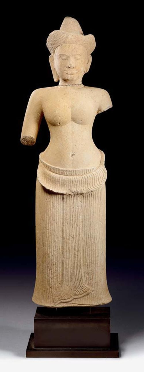 FEMALE DEITY.Khmer, in the style of Angkor Vat, 12th century. H 72 cm. Sandstone. Classic depiction with a well-formed upper body and a skirt with pleated vertical folds, which is held together by a patterned belt. On the front: the collar trails beneath the belt, the overlap ends in a point shaped like a fish-tail. An engraved crown adorns the plaited hair-do. Gentle and friendly facial expression. Head attached.