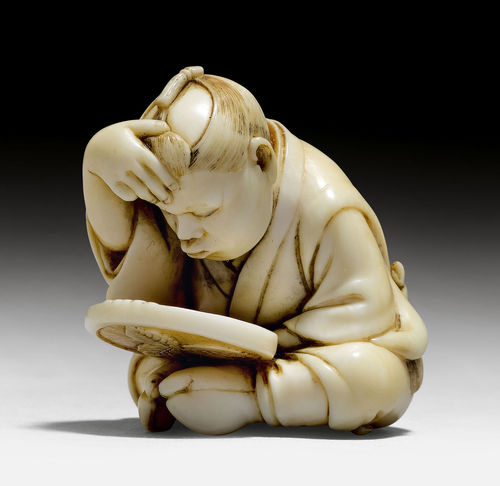 AN IVORY NETSUKE OF A BOY EXAMINING HIMSELF IN A MIRROR.