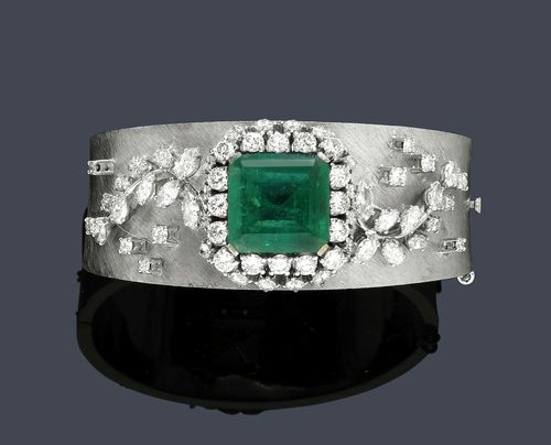 EMERALD AND DIAMOND BANGLE, ca. 1950. White gold ca. 700, 99g. Broad satin-finished bangle with hinge, set with 1 step-cut Columbian emerald of ca. 14.00 ct, minimal signs of wear, within a double-border of 32 brilliant-cut diamonds weighing ca. 3.50 ct and additionally decorated with 2 appliqued leaf motifs set with 22 navette-cut diamonds and 10 brilliant-cut diamonds. Total diamond weight 2.30 ct. W ca. 2.2 cm, ca. 6 x 5 cm. Tested by Gemlab.