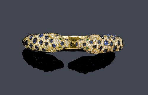 SAPPHIRE, DIAMOND AND EMERALD CLASP. Yellow gold 750. Very attractive gold bangle with  hinge, the ends designed as panther heads, set throughout with numerous sapphire cabochons weighing ca. 20.00 ct. and numerous single-cut diamonds weighing ca. 6.00 ct, some missing. Four small emeralds as eyes. Ca. 4.5 x 6 cm.