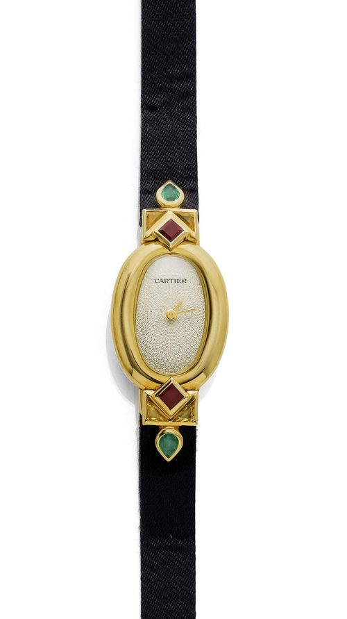 RUBY AND EMERALD LADY'S WRISTWATCH, CARTIER BAIGNOIRE, 1990s. Yellow gold 750. "Mini-Baignoire" model. Oval, slightly convex case No. GC10127, with screw-down back, attaches set with 2 square-cut rubies, 4 triangle-cut sapphires and 2 drop-cut emeralds weighing ca. 1.00 ct. Fine, engine-turned dial with gold-coloured hands. Quartz movement. Black satin band with fold-over clasp. D 34 x 18 mm. With case, warranty, and setting tool, November 1994.