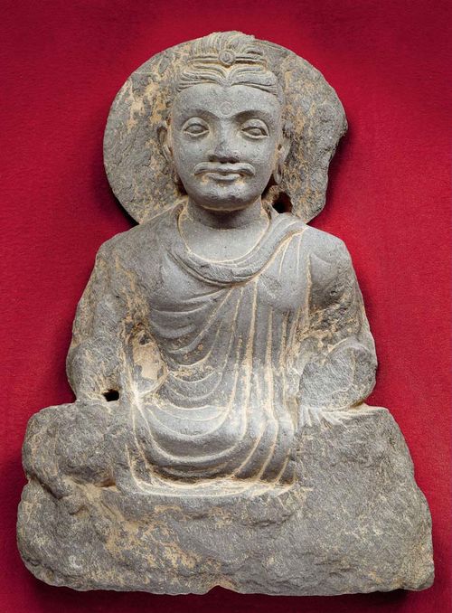 SITTING BUDDHA.Gandhara, 2nd/3rd century. H 36 cm. Grey slate. The head in front of a lotus aureole is almost plastically round, the body is worked in half relief. A simple monk's robe with even pleats is draped over the body.