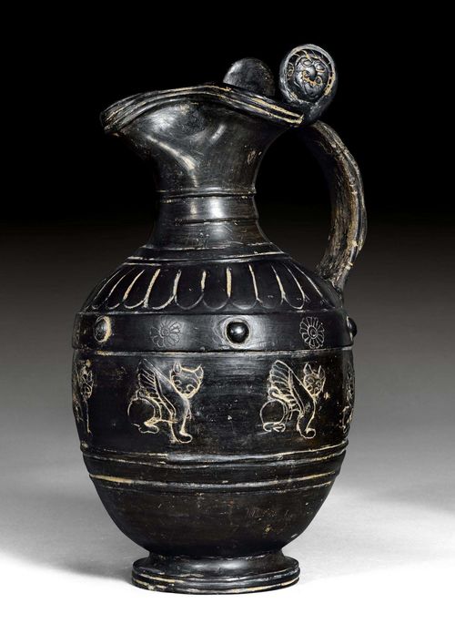 JUG WITH HANDLE,so-called "Oinorche", Etruria, 2nd half of the 6th century BC. Black painted ceramics. Jug with a reliefed handle and 2 stylised faces as well as with a curved spout, on a rounded base. Walls with a stylized sphinx decor. H 36.4 cm. Provenance: Austrian private collection, acquired in Vienna in the 1980s.