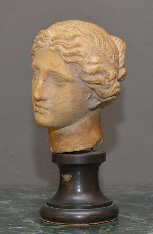 HEAD OF A NYMPH,Roman, after a Hellenistic example from the 2nd century BC, Italy, 1st/2nd century AD. Beige stone. Fine head with wavy hair with a bun, on a conical, round wooden base with applied crown. H 16.5 cm (without base). H 24 cm (with base).