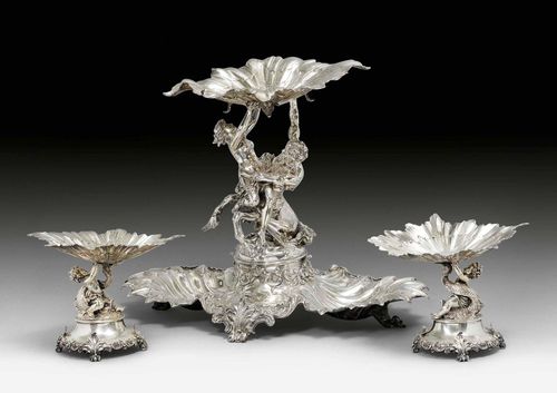 DINNER SERVICE, Berlin, end of the 19th century. Maker's mark: Paul Telge. Imposing centrepiece, the middle with two intertwined Gods holding a bowl with their outstretched arms. Feet of the bowls, later, probably so as to use them separately. Two matching side pieces. Oval base on four volute feet. H 52 cm, 7745g.