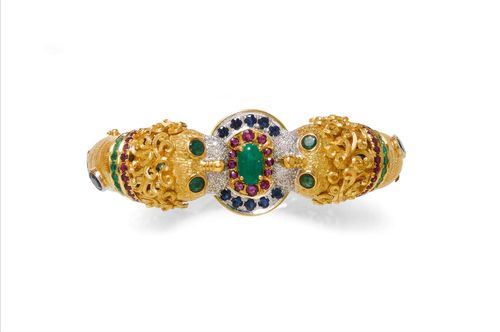 GEMSTONE AND DIAMOND BANGLE, ca. 1960. Yellow gold 750, 77g. Decorative bangle with hinge and a textured surface, set with 9 oval sapphires. The ends designed as 2 lion heads set with diamonds and with a collar of gemstones, in-between: a ring set throughout with gemstones. Total weight of the emeralds ca. 4.30 ct, of the sapphires ca. 10.40 ct, of the rubies ca. 3.00 ct, and of the diamonds ca. 0.60 ct.