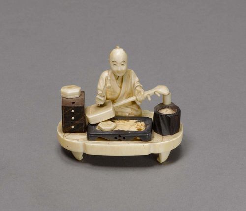 OKIMONO OF A MUSICAL INSTRUMENT MAKER.Japan, Meiji Period, L 7.5 cm. Ivory and wood. The seated man holds a hammer in his right hand and is working on a shamisen. Light dam. In addition: an arm with an open book, which one can insert before the craftsmen but probably did not belong to it originally.