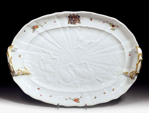OVAL PLATTER WITH REED HANDLES FROM THE SWAN SERVICE, Meissen, circa 1740.Model by Johann Friedrich Eberlein, 1740. The well relief-decorated with two swans in the reeds, the ribbed sides with shell decoration, painted with small Indianischen Blumen and crowned with the Brühl-Kolowrat-Krakowsky coat of arms. Underglaze blue sword mark, impressed number 27. D 38.3cm. The gilding on the edge missing. The gilding on the handles of a later date.