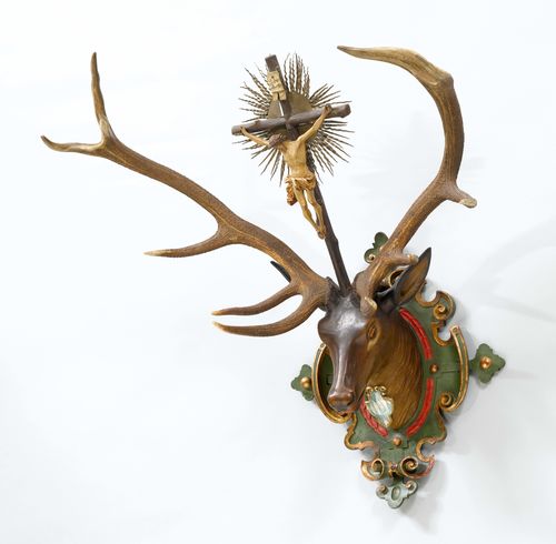 DEER HEAD WITH ANTLERS, 19th century. Wood, carved and painted green/red/gold, with crucifix between the antlers. Bavarian coat-of-arms. 90 x 114 cm.