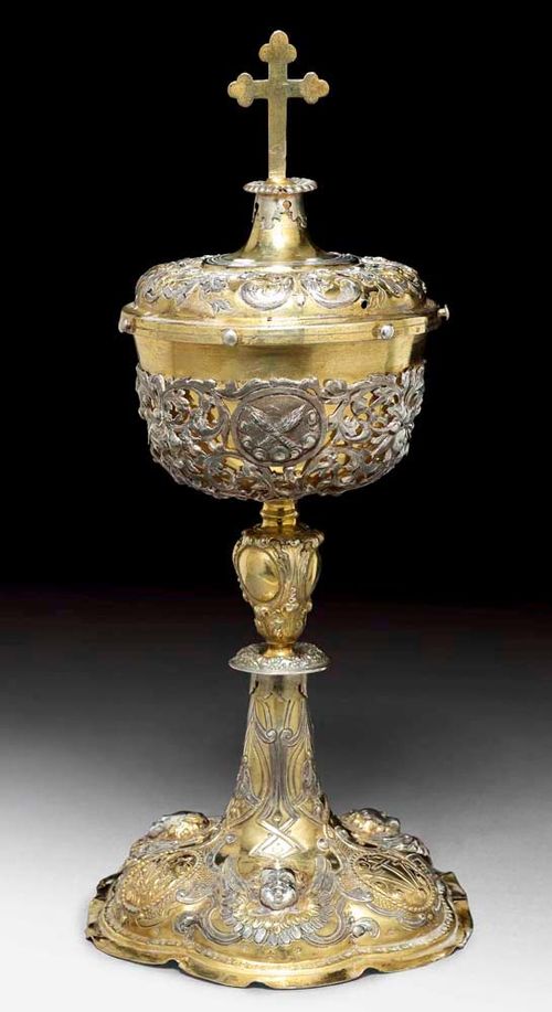 ZIBORIUM. Augsburg, 1708-10.Maker's mark  Ludwig Schneider. Parcel gilt. With chased and embossed decoration of angels' heads and scrolls. The cup with openwork silver mount. The domed lid with acanthus, flowers and cross as finial. H 36cm, 750 g.