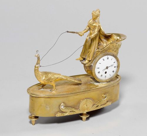 SMALL TABLE CLOCK,Empire, probably France. The movement signed PATRON À GENÈVE. Gilt bronze. Designed as a woman on a chariot drawn by a peacock. On an oval plinth with spherical feet. White enamel dial (chipped). 21x8x22 cm. 2 feet missing, requires restoration.