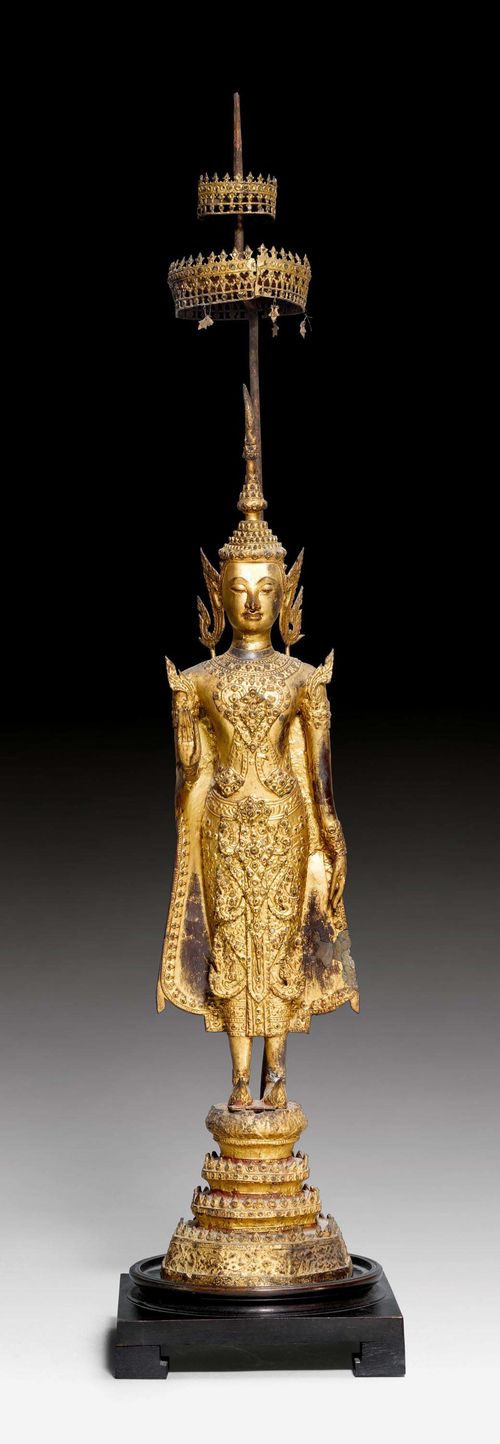 A GILT AND LACQUERED BRONZE FIGURE OF A STANDING BUDDHA WITH PARASOL. Thailand, 19th c. 60 cm (without parasol). Painted in parts. Brittle. Added wood base.