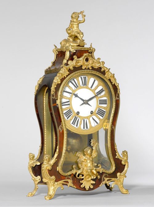 TABLE CLOCK,in the Regence style, France, 19th century. Curved, wooden case, decorated with red tortoiseshell. Bronze mounts designed as rocailles, flowers and heads. Sculpted putto on top.  Bronze dial with enamel cartouches. Movement with verge escapement, striking the 1/2-hour on bell. H 57 cm. Losses.
