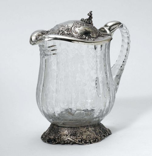 WATER PITCHER WITH SILVER MOUNT,Germany, after 1888. Oval, curved stand decorated with volutes. Glass vessel with cut grape-vines and volutes. Hinged lid with volute cartouches. H 26 cm.