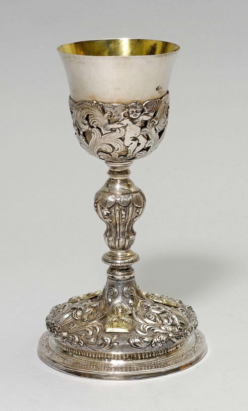 ECCLESIASTICAL CUP, probably 19th century. In the Baroque style. Convex, chased and chiselled round foot. Decorated with saints and applied plaques with saints. Gilt cuppa with winged putti and pierced volutes. H 24 cm.
