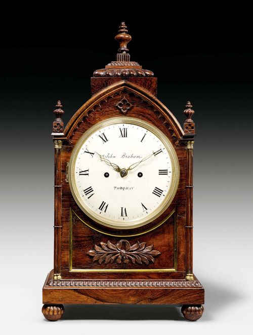 CLOCK, Gothic Revival, England, mid-19th century. The movement and dial signed JOHN BRAHAM TORQAY. Rosewood. Brass fillets and capitals. White painted metal dial. Anchor escapement striking the ½-hours on gong. Repetition on demand. H 54 cm.