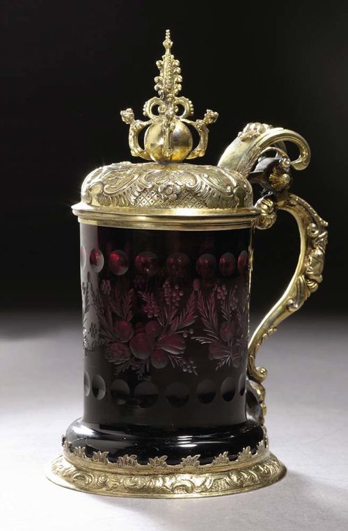 SILVER GILT TANKARD AND COVER WITH RUBY RED GLASS 19/20th century. In the style of the 17th century. No mark. Chased base ring with scrolls and rocaille. The lid with 3 images between rocaille and flowers, ball finial with female figures, the handle in the form of a face. The glass with cut floral ornament. H 20 cm.