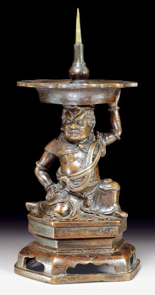 CANDLESTICK.China, Ming Dynasty, H 28 cm. Bronze. A curly-haired, frowning guard figurine with a shawl draped over his shoulders and scarves wrapped around his torso is kneeling on a hexagonal pedestal. One of the guard's hands is supporting the curved bowl, which he is balancing on his head, the other hand is resting on his thigh.