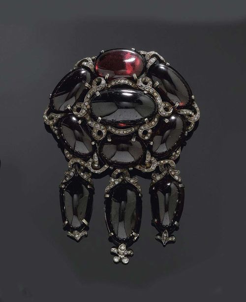 GARNET AND DIAMOND BROOCH, circa 1890. Silver, parcel gilt. The oval top set with 7 large oval garnet cabochons of ca. 24.5x13.7 to 17.5 x 11mm, with fine band motifs set with small diamond roses. The terminal consisting of 3 drop shaped garnet cabochons on moveable mount, ca. 21.2x13.5 to 19x11.8mm, and decorated with stylised floral and foliate motifs set with diamond roses. Brooch pin can be screwed off. Small repairs.