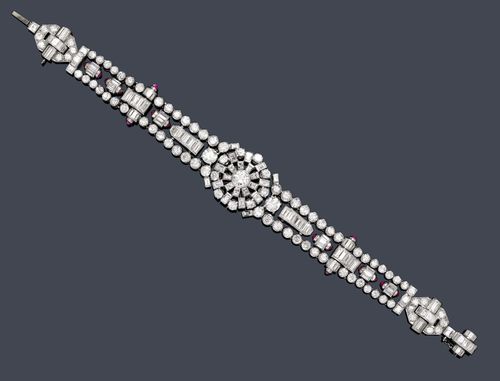 DIAMOND BRACELET, probably France, ca. 1930. Platinum. Elegant, geometrically designed Art Deco bracelet, the round centre part decorated in a ray-like manner with numerous baguette-cut diamonds and old European-cut diamonds, the sides of 2 lines of brilliant-cut diamonds and, in-between, differently combined cylinder motifs with baguette-cut diamonds and small ruby cabochons as well as diamond-set band motifs. Total weight of the ca. 97 baguette-cut diamonds and the 72 old European-cut diamonds ca. 10.00 ct. L ca. 18 cm. With case signed Fontana F.rs, Galerie des Valois 122,123,124, Palais Royal Paris.