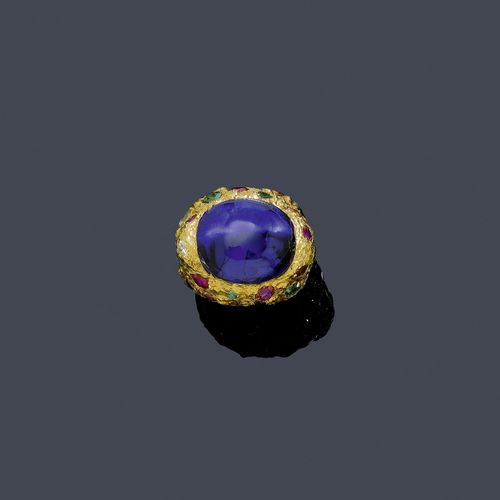 TANZANITE, GEMSTONE AND DIAMOND RING. Yellow gold 750. Casual-elegant mantle ring with a textured surface, the top set with 1 fine, blue oval tanzanite cabochon of ca. 28.00 ct decorated with 10 tourmalines, 8 rubies, 2 sapphires, 2 peridots, 2 citrines, 1 emerald and 1 brilliant-cut diamond weighing ca. 4.00 ct. Size ca. 50. With report by Walter Grün, October 2003.