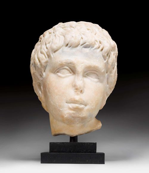 HEAD OF EROS, Roman, according to the Eros by Lysippos, Italy, 1st century AD. Beige marble. Mounted on a black, stepped wooden base. Chipped. Head: H 22.5 cm. With base: H 28 cm. Provenance: - in a Swiss private collection since 1974. A notarized document is available at Galerie Koller. With opinion by Dr. J.D. Chamay, Geneva, 15 June 2014.