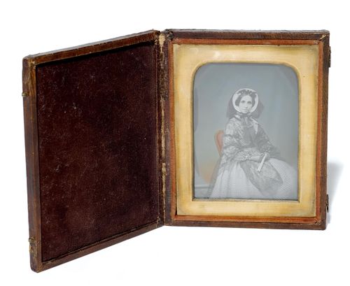 AMBROTYPIEN - [Anonym]. Portrait einer Frau. Kol. Original-Photographie. Ambrotypie. Wohl England, um 1860. Abgerundeter Bildausschnitt, ca. 8,8 x 6,5 cm. Alt unter Glas mont. mit Messing-Passepartout in Holz-Kassette mit Lederbezug u. 2 intakten Schliessen, Stoffeinlage (ca. 10 x 12,6x 2 cm; minimal berieben).