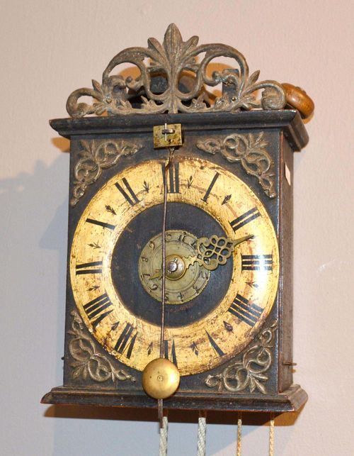 WALL CLOCK WITH ALARM AND FRONT PENDULUM,Baroque, Switzerland. Blackened wood with open-worked tin ornament. Fronton with wood chapter ring and alarm disc. Verge escapement. Alarm on bell. H 27 cm.