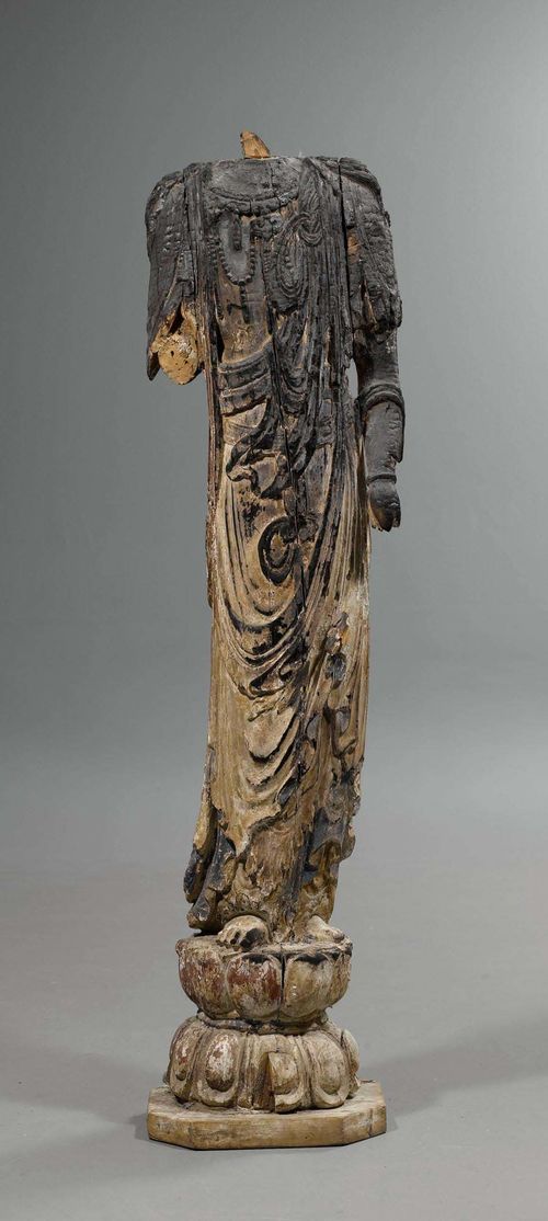 STANDING BUDDHA.China, Ming dynasty or earlier, H 165 cm. Wood with traces of polychrome paint. The figure with finely sculpted robe and jewellery stands on a double lotus pedestal. Despite burn marks the high quality of the carving is still evident. The head, the right forearm, and the left hand are lost.