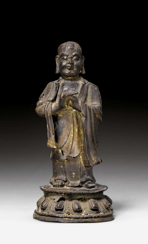 STANDING KASYAPA.China, Ming dynasty, H 20.5 cm. Bronze. In long robes, which leave his chest and right arm bare, the monk stands barefoot on a lotus pedestal. His hands are folded before his body. He is among the favourite disciples of the Buddha.