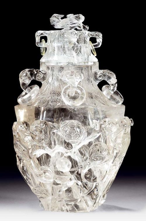 ROCK CRYSTAL VESSEL.China, 19th/20th century. H 32 cm. Very large vessel with lid in the shape of a hexagonal vase with four ring handles on its neck. The lower part of the vase has carvings of two birds between rampant flowers. The neck and the lid have banana leaf wreaths in flat relief. The lid is crowned by a bird holding a flower in its beak. Two small rings have been added.
